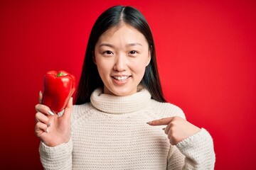 Young asian woman holding healthy and fresh red pepper over isolated background with surprise face pointing finger to himself