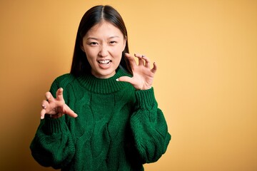 Young beautiful asian woman wearing green winter sweater over yellow isolated background smiling funny doing claw gesture as cat, aggressive and sexy expression