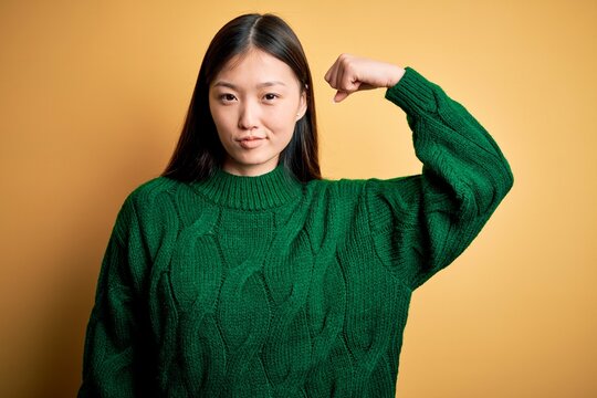Young beautiful asian woman wearing green winter sweater over yellow isolated background Strong person showing arm muscle, confident and proud of power