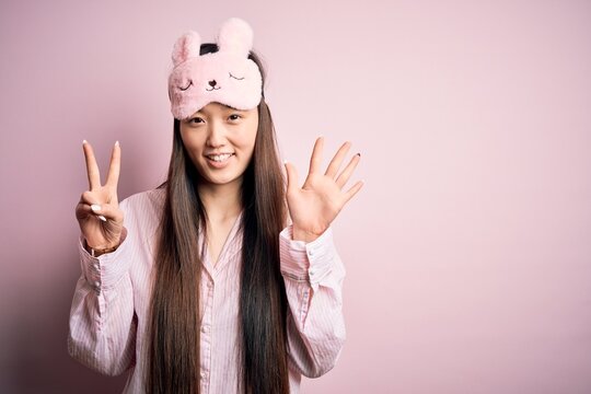 Young Asian Woman Wearing Pajama And Sleep Mask Over Pink Isolated Background Showing And Pointing Up With Fingers Number Seven While Smiling Confident And Happy.