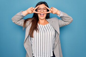 Young hispanic business woman wearing glasses standing over blue isolated background Doing peace symbol with fingers over face, smiling cheerful showing victory