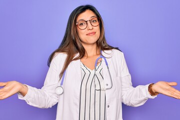 Professional doctor woman wearing stethoscope and medical coat over purple background clueless and...