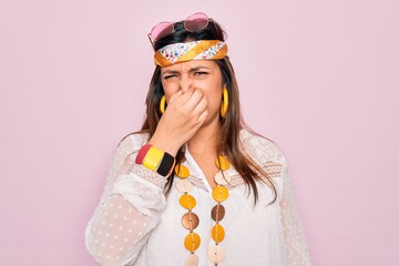 Young hispanic hippie woman wearing fashion boho style and sunglasses over pink background smelling something stinky and disgusting, intolerable smell, holding breath with fingers on nose. Bad smell