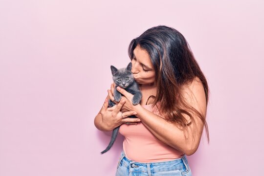 Young beautiful latin woman smilling happy. Standing with smile on face holding and kissing adorable cat over isolated pink background