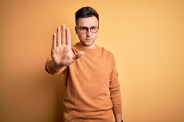 Young handsome caucasian man wearing glasses and casual winter sweater over yellow background doing stop sing with palm of the hand. Warning expression with negative and serious gesture on the face.