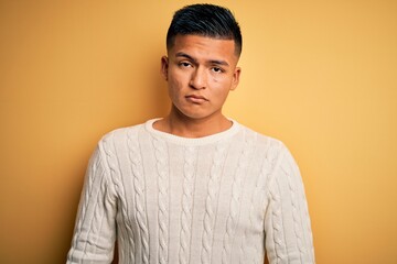 Young handsome latin man wearing white casual sweater over yellow background with serious expression on face. Simple and natural looking at the camera.