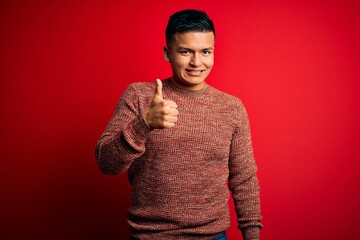 Young handsome latin man wearing casual sweater standing over red background doing happy thumbs up gesture with hand. Approving expression looking at the camera showing success.