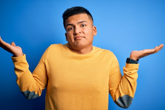 Young handsome latin man wearing yellow casual sweater over isolated blue background clueless and confused expression with arms and hands raised. Doubt concept.
