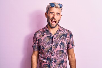 Handsome blond man on vacation wearing casual shirt and sunglasses over pink background winking looking at the camera with sexy expression, cheerful and happy face.