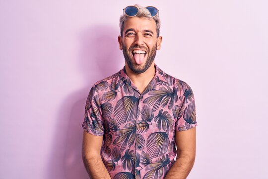 Handsome blond man on vacation wearing casual shirt and sunglasses over pink background sticking tongue out happy with funny expression. Emotion concept.