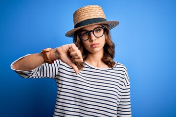 Young beautiful blonde girl wearing glasses and hat over blue isolated background with angry face, negative sign showing dislike with thumbs down, rejection concept