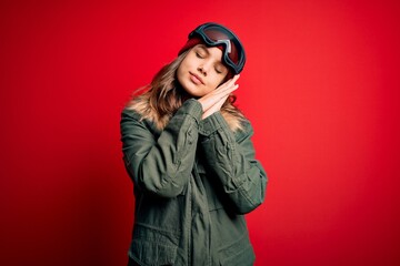 Young blonde girl wearing ski glasses and winter coat for ski weather over red background sleeping tired dreaming and posing with hands together while smiling with closed eyes.