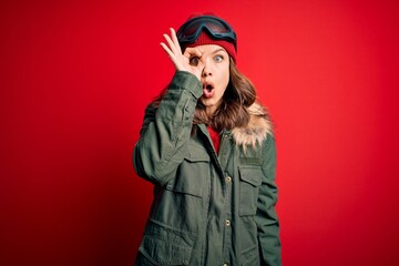 Young blonde girl wearing ski glasses and winter coat for ski weather over red background doing ok gesture shocked with surprised face, eye looking through fingers. Unbelieving expression.