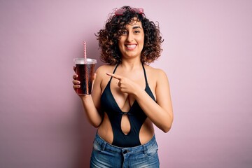 Beautiful arab woman on vacation wearing swimsuit drinking cola refreshment using straw very happy pointing with hand and finger