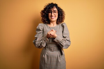 Beautiful arab business woman wearing dress and glasses standing over yellow background with hands together and crossed fingers smiling relaxed and cheerful. Success and optimistic