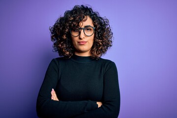 Young beautiful curly arab woman wearing casual sweater and glasses over purple background skeptic and nervous, disapproving expression on face with crossed arms. Negative person.
