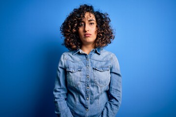 Young beautiful curly arab woman wearing casual denim shirt standing over blue background Relaxed with serious expression on face. Simple and natural looking at the camera.