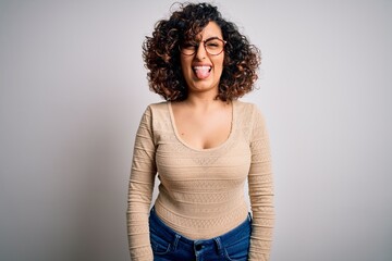 Young beautiful curly arab woman wearing casual t-shirt and glasses over white background sticking tongue out happy with funny expression. Emotion concept.