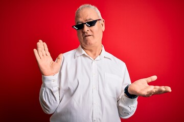 Middle age hoary man wearing funny sunglasses over isolated red background clueless and confused expression with arms and hands raised. Doubt concept.