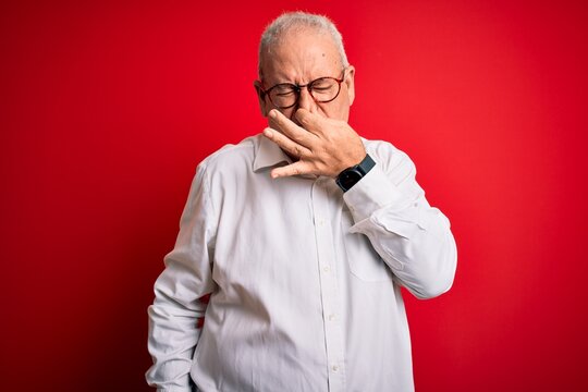 Middle age handsome hoary man wearing casual shirt and glasses over red background smelling something stinky and disgusting, intolerable smell, holding breath with fingers on nose. Bad smell