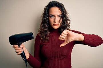 Young beautiful woman with curly hair using hair dryer over isolated white background with angry...