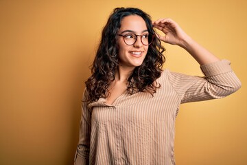 Beautiful woman with curly hair wearing striped shirt and glasses over yellow background smiling confident touching hair with hand up gesture, posing attractive and fashionable