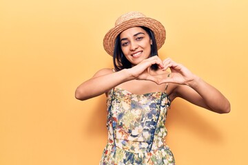 Young beautiful latin woman wearing summer hat smiling in love doing heart symbol shape with hands. romantic concept.