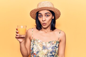 Young beautiful latin woman wearing summer hat drinking glass of orange juice scared and amazed with open mouth for surprise, disbelief face