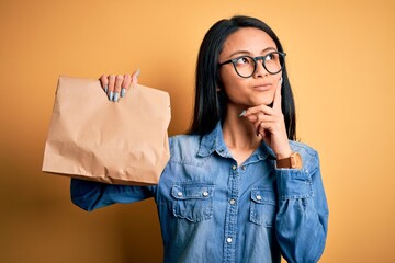 Young beautiful chinese woman holding delivery bag over isolated yellow background serious face...