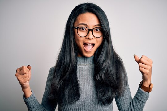 Young beautiful chinese woman wearing glasses and sweater over isolated white background celebrating surprised and amazed for success with arms raised and open eyes. Winner concept.