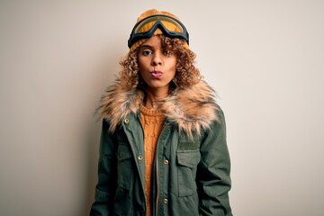 Young african american skier woman with curly hair wearing snow sportswear and ski goggles looking at the camera blowing a kiss on air being lovely and sexy. Love expression.