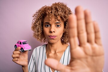 Young african american woman holding small pink car standing over isolated pink background with open hand doing stop sign with serious and confident expression, defense gesture