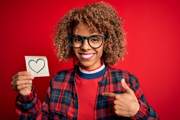 Young african american curly woman holding reminder paper with heart over red background with surprise face pointing finger to himself