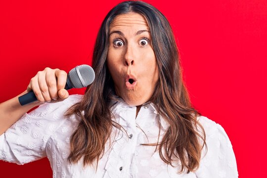 Young beautiful brunette singer woman singing song using microphone over red background scared and amazed with open mouth for surprise, disbelief face