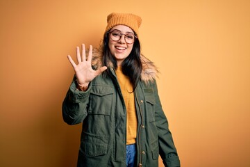 Young brunette woman wearing glasses and winter coat with hat over yellow isolated background...