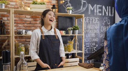 Laughing asian japanese female barista cheerful chatting with muslim lady client in cafe bar counter. girl bartender in apron greeting friend regular customer in coffeehouse. nice service concept