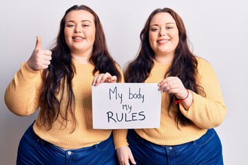Young plus size twins holding my body my rules banner smiling happy and positive, thumb up doing excellent and approval sign