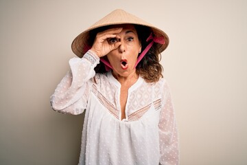 Middle age brunette woman wearing asian traditional conical hat over white background doing ok gesture shocked with surprised face, eye looking through fingers. Unbelieving expression.