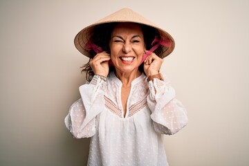 Middle age brunette woman wearing asian traditional conical hat over white background covering ears with fingers with annoyed expression for the noise of loud music. Deaf concept.