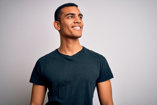 Young handsome african american man wearing casual t-shirt standing over white background looking away to side with smile on face, natural expression. Laughing confident.