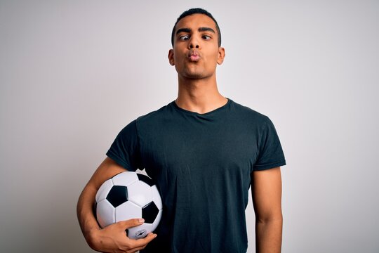 Handsome African American Man Playing Footbal Holding Soccer Ball Over White Background Making Fish Face With Lips, Crazy And Comical Gesture. Funny Expression.