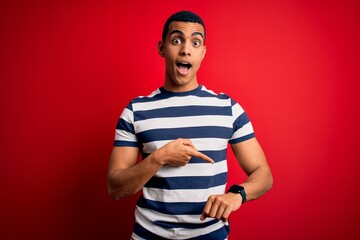 Handsome african american man wearing casual striped t-shirt standing over red background In hurry pointing to watch time, impatience, upset and angry for deadline delay