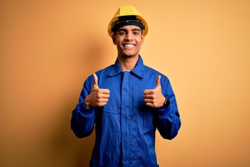 Young handsome african american worker man wearing blue uniform and security helmet success sign doing positive gesture with hand, thumbs up smiling and happy. Cheerful expression and winner gesture.