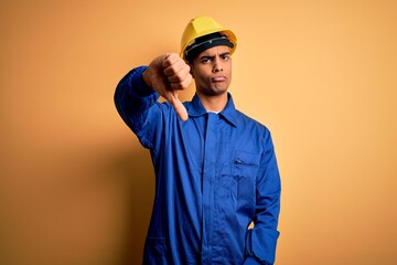 Young handsome african american worker man wearing blue uniform and security helmet looking unhappy and angry showing rejection and negative with thumbs down gesture. Bad expression.