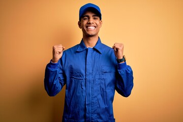 Young african american mechanic man wearing blue uniform and cap over yellow background celebrating...