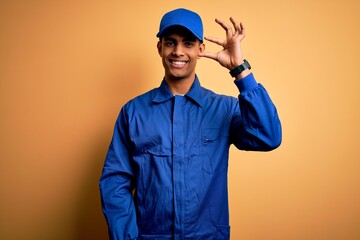 Young african american mechanic man wearing blue uniform and cap over yellow background smiling and confident gesturing with hand doing small size sign with fingers looking and the camera. Measure