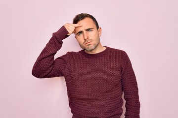Young handsome man with blue eyes wearing casual sweater standing over pink background pointing unhappy to pimple on forehead, ugly infection of blackhead. Acne and skin problem