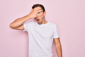 Handsome man with blue eyes wearing casual white t-shirt standing over pink background peeking in shock covering face and eyes with hand, looking through fingers with embarrassed expression.