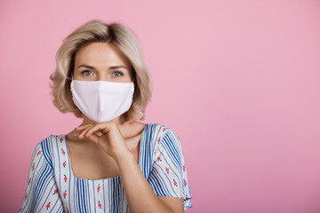 Blonde woman with a medical mask on face looking at camera while wearing a dress and touching her chin on a pink studio wall with free space