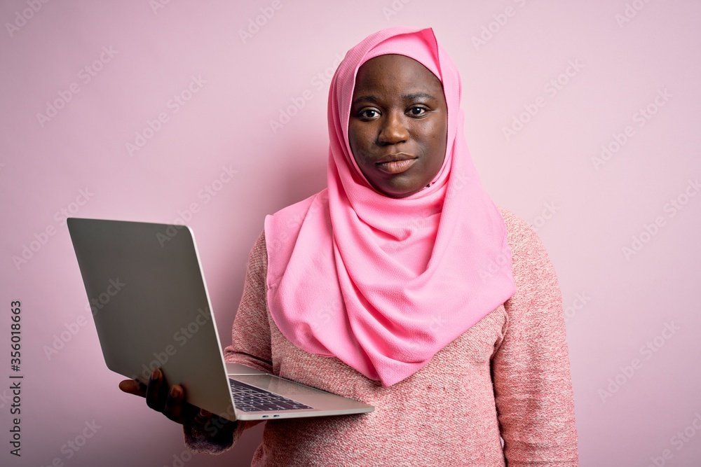 Sticker African american plus size woman wearing muslim hijab using laptop over pink background Relaxed with serious expression on face. Simple and natural looking at the camera.